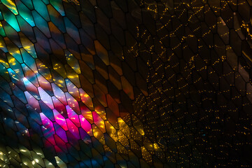 abstract, geometric wall made of hexagonal, glass-like tiles. The tiles reflect colorful lights creating a vibrant, shimmering effect. The overall texture is sleek and modern, almost cosmic aesthetic