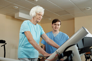 Elderly Caucasian woman walking on treadmill under control of nursing assistant while getting through physical therapy in rehab hospital
