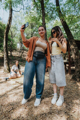 Two young women pose for a selfie in a leafy forest while a friend relaxes on a blanket nearby. Warm, casual outdoor moment suitable for lifestyle, friendship, and summer content.