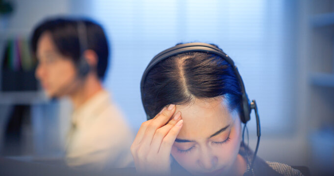Tired Asian call center employee wearing headset touching forehead from fatigue while working in office at night, stress and burnout concept