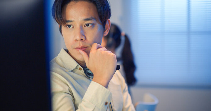 Serious Asian man with headset focusing on computer screen, analyzing data and working in modern office at night.
