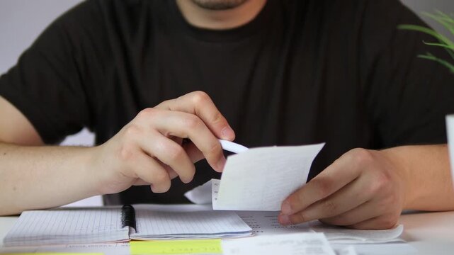 A man's hands at a home desk carefully cross-reference a paper receipt with an open notebook, using a pen to verify details. 