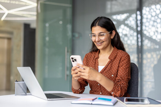 Young indian woman in glasses sitting at her office desk, happily interacting with a smartphone while a laptop and documents are nearby, representing modern business communication and technology use - Powered by Adobe