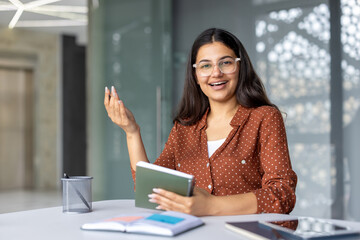 Young indian businesswoman in glasses and polka-dot shirt smiling and speaking at a meeting,...