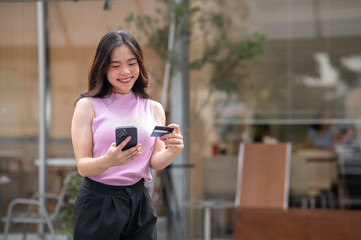 Asian young woman holding credit card looking at phone with smile while standing in front of a cafe.