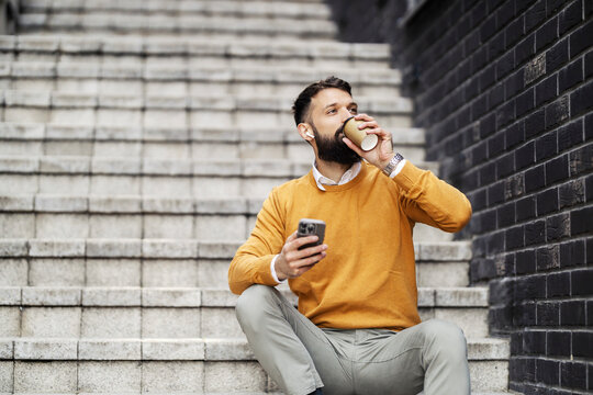 Portrait of businessman relaxing on a staircase with cellphone in hands and refreshing with coffee on a coffee break.