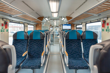 Wide train view. Rows of patterned blue train seats line both sides of the carriage. Overhead racks and clear windows give the space a bright look.