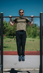 Obraz premium A man showcases his strength and fitness by doing pull-ups at an outdoor exercise park on a sunny day with green scenery.