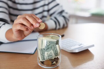 Woman with calculator putting money into jar at wooden table indoors, closeup