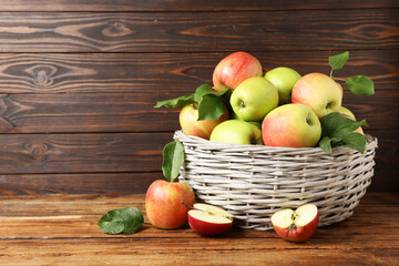 Fresh ripe apples with green leaves in wicker basket on wooden table, space for text