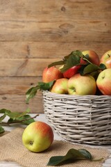 Fresh ripe apples with green leaves in wicker basket on white wooden table, closeup