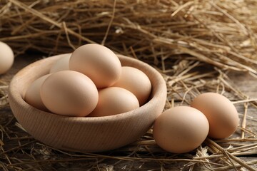 Raw chicken eggs in bowl and straw on wooden table, closeup