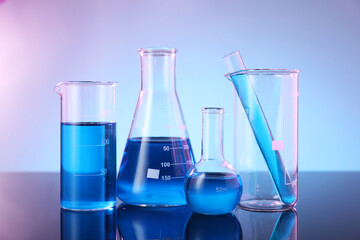 Different glassware with blue liquid on mirror surface against color background