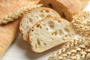 Fresh crispy ciabattas and wheat ears on white wooden table, closeup