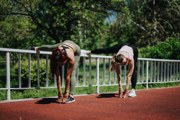 Two women in active wear warm up by stretching near a railing on a sunny day outdoors. Ideal image...