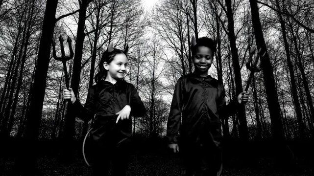 Devil Costumes Kids Forest Black and White - Two children, a girl and a boy, dressed in devil costumes with pitchforks, playfully interact in a dark forest setting.