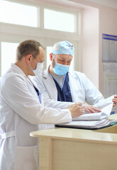 Two male doctors in protective medical masks discussing patient diagnosis and reviewing medical documents in a hospital. Concept of healthcare teamwork, professional medicine, and hospital routine.