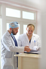 Two male doctors in protective medical masks discussing patient diagnosis and reviewing medical documents in a hospital. Concept of healthcare teamwork, professional medicine, and hospital routine.