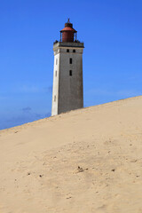 Photo with a view of the Rubjerg Knude lighthouse (also known as the "Lighthouse in the Dunes") with sand dunes in the foreground against a blue sky with clouds near Lokken in Denmark