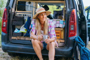 Sitting by her minivan with coffee in hand a woman relaxes after a lakeside hike enjoying wild camping life