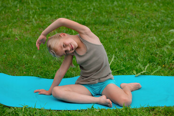 A 9-year-old blonde girl does exercises on a mat outdoors.
