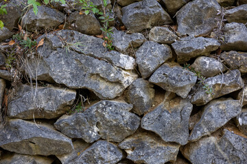 Old stone wall made of irregular rocks with plants growing between, reflecting rustic architecture, natural materials and countryside heritage