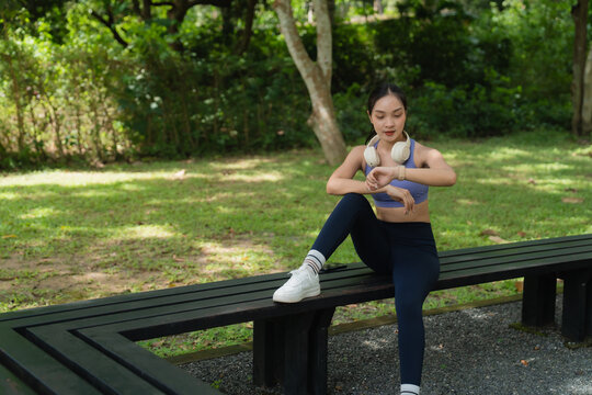 Young Asian woman taking a break from workout in an outdoor park, sitting on a bench while looking at her smartwatch, tracking progress and monitoring fitness activity