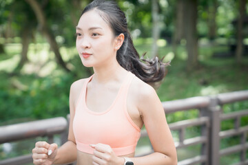 Asian woman jogging through a lush green park in an orange sports bra, focused on fitness and...