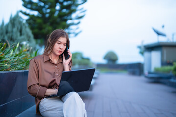 Young asian businesswoman sitting on a bench in an outdoor urban setting, multitasking between a laptop and a smartphone, demonstrating remote work and digital nomad lifestyle