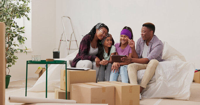 A married couple with their two daughters, an African American family, sits on the couch in their apartment. They browse a tablet, seeking inspiration for completing the rooms and deciding on paint