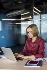Obraz premium Professional businesswoman wearing glasses and a formal shirt typing on a laptop computer, managing tasks at a wooden desk in a contemporary, glass-walled office environment