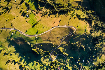 Top-down aerial view of picturesque rural landscape bathed in golden sunlight. Winding roads meander through green fields and small patches of trees. Scattered farmhouses and haystacks dot terrain.