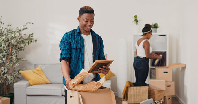 A man unpacks cardboard boxes in their new apartment. He finds an old photo frame and shows it to his wife. They both smile and laugh, reminiscing about the happy memories of the past.
