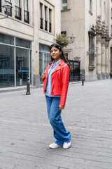 A teenager in a red jacket and blue jeans stands on a sidewalk