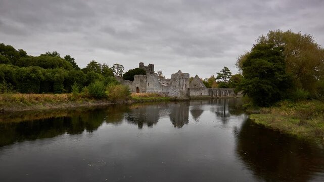 Desmond Castle in Adare, Ireland, reflected in a tranquil river surrounded by lush greenery and a cloudy sky