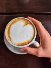 Hand holding a cup of coffee with heart-shaped foam design on a wooden table