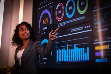 Hispanic latin business woman interacting with virtual data interface. African american female observes financial data displayed on a large screen monitor, contemplating market trends and strategy.