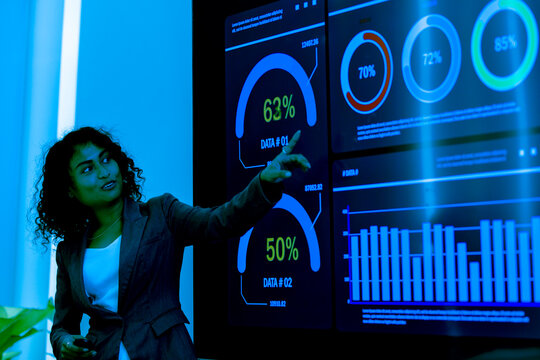 Hispanic latin business woman interacting with virtual data interface. African american female observes financial data displayed on a large screen monitor, contemplating market trends and strategy.