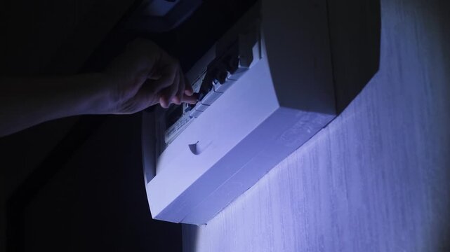 Man hand turns on circuit breaker in electrical panel in dark room with flashlight light home lighting repair electrical equipment