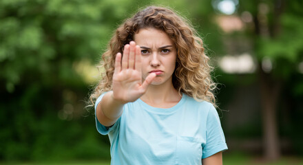 Fototapeta premium Young woman with curly hair making a stop gesture with her hand, conveying a clear message of refusal or boundary setting outdoors