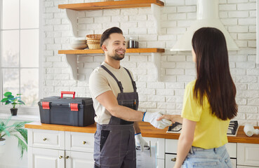 Smiling young handyman dressed in overalls shakes hands with female client, standing together in...