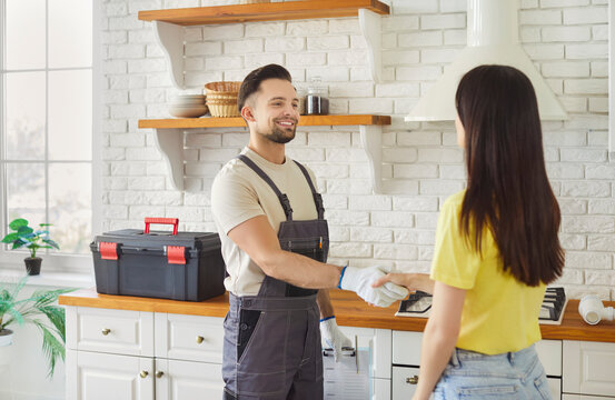 Smiling young handyman dressed in overalls shakes hands with female client, standing together in modern kitchen. Making agreement, successful completion of home repair, professional services concept