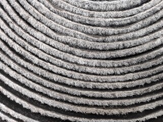 Gray striped background, rolled-up carpet. Close-up view of textured concentric circles with a rough, fibrous appearance. The pattern creates a hypnotic and repetitive visual effect.