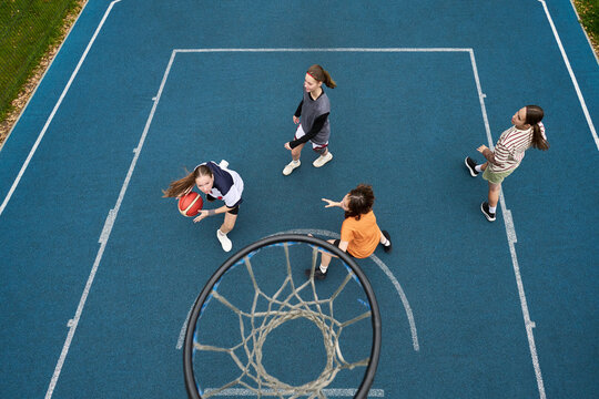 Group of multiethnic teenagers playing streetball on outdoor court, Caucasian girl dribbling basketball while other teenagers defending and observing, hoop visible in foreground