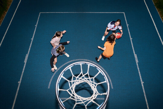 Group of four teenage girls playing streetball on outdoor court, two defending while one dribbling basketball and another observing, photographed from above through basketball hoop