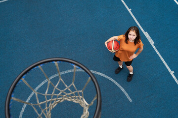 Portrait of Caucasian teenage girl standing on outdoor basketball court holding basketball looking up toward camera near hoop representing streetball concept