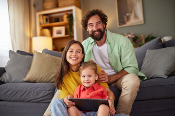 In a warm living room, a smiling family sits together on a comfortable couch. Parents hold their child while watching something entertaining on a tablet, creating joyful moments together.