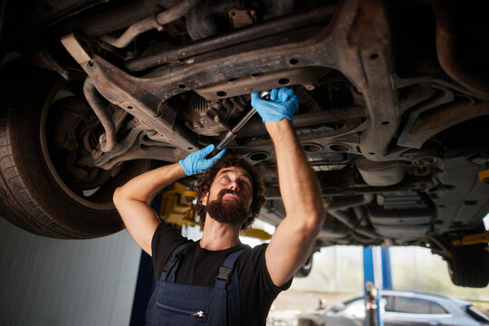 A skilled car mechanic is working underneath a vehicle in a busy workshop, using a wrench to tighten components. The area is well-lit, showing tools and equipment around.
