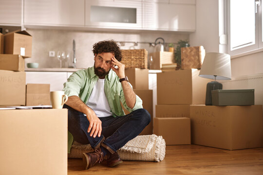 A man sits on the floor surrounded by unpacked boxes in a new home, looking pensive and overwhelmed as he contemplates the moving process and settling in.