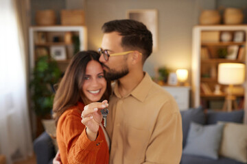 Couple joyfully poses together in their new house, holding keys and sharing a kiss in a bright, stylish living room that exudes warmth and comfort.